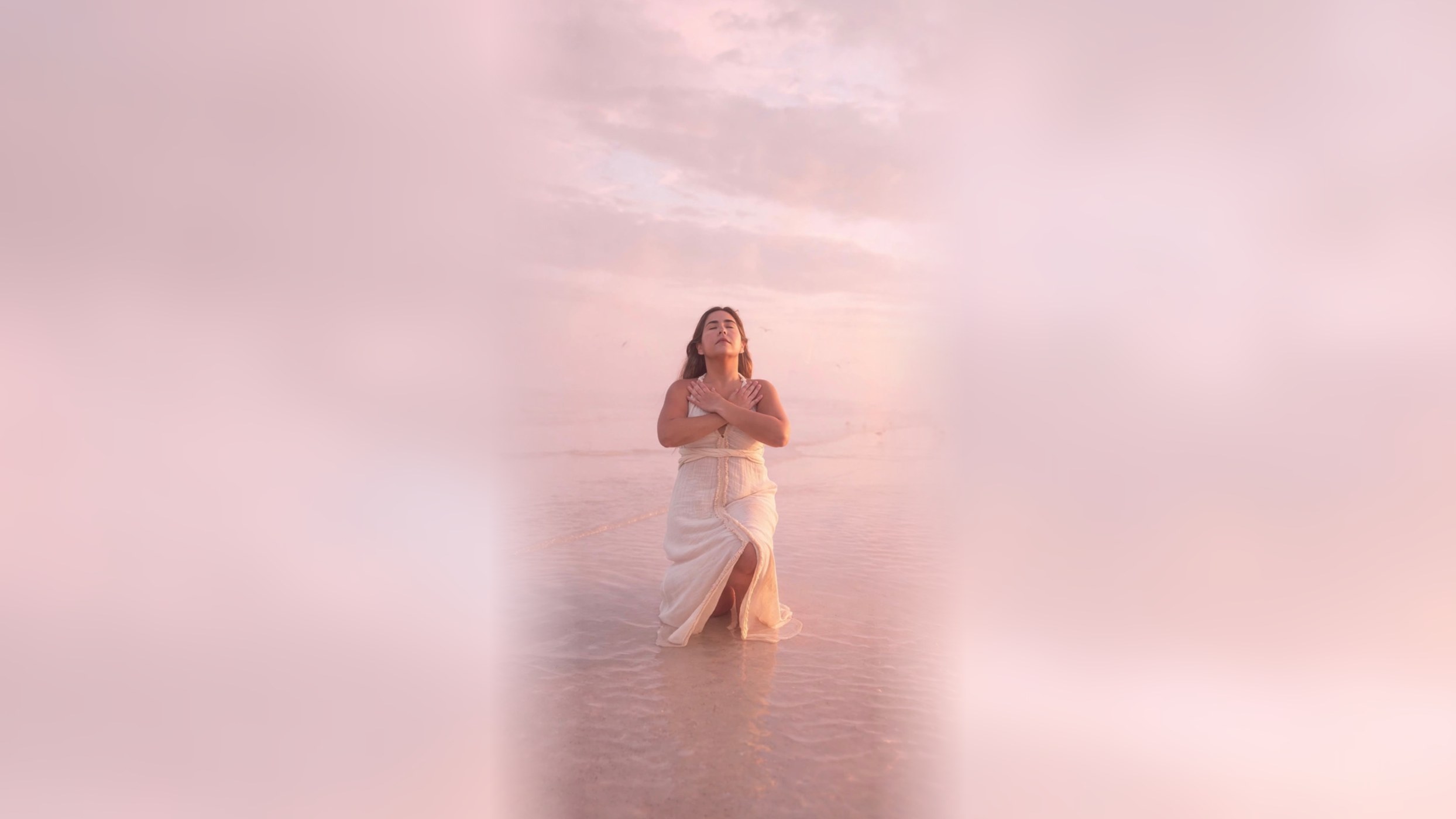 Celeste kneeling in shallow pink ocean water at sunset, hands crossed over her heart